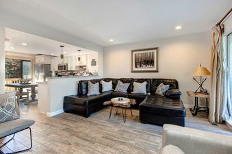 Living room with a sectional sofa, a large window with a view of the outside, and a kitchen in the background.
