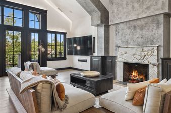 A modern living room at The Gallatin with a vaulted ceiling, large windows overlooking trees, a fireplace with a marble surround, a large TV, and comfortable sofas.
