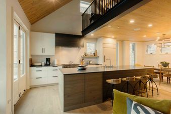 The kitchen at Transfer Haus features white cabinetry, a dark gray countertop, and a large island with wooden stools, complemented by a light wood ceiling and an upper-level balcony.