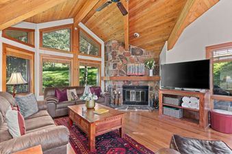 A spacious living room at Crescent Cabin with a large stone fireplace, brown leather sofas, and numerous windows offering outdoor views.
