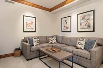 The living room in Grand Lodge Three Bedroom Deluxe 408 features a sectional couch with a coffee table, and three vintage ski posters on the walls.