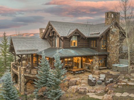 A luxurious mountain home, High Road, featuring a stone and timber exterior, large decks, a hot tub, and landscaped gardens under a dramatic sunset sky.