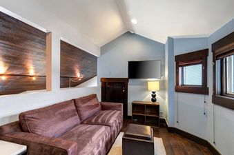 A cozy living room with a purple sofa, a mounted TV, a lamp on a wooden shelf, and windows with dark wood frames.