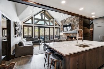 A spacious, open-concept living area and kitchen in Snow Cloud Lodge 506, featuring a large stone fireplace, vaulted ceilings, and floor-to-ceiling windows that offer scenic outdoor views.
