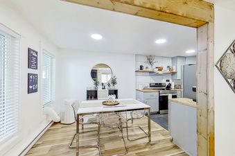 A white dining table with clear chairs is set in a modern kitchen with white walls, hardwood floors, and a view of the kitchen counter with stainless steel appliances.