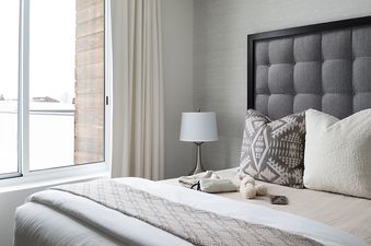 A cozy bedroom at Penthouse on Glenwood 407 featuring a tufted gray headboard, elegant bedding with patterned and boucle pillows, and a bedside lamp, all set against a textured wallpaper, with a large window revealing a snowy outdoor scene.