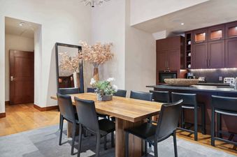 A dining room with a large wooden table and black leather chairs. There are plants and a mirror in the background, and a kitchen with a microwave and coffee maker are visible in the back of the room.