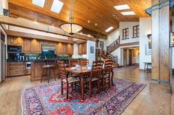 The Peaks Penthouse 702 showcases a spacious dining area with a wooden table and chairs, adjacent to an open-plan kitchen, all under a vaulted wooden ceiling with skylights.