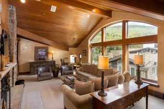 A spacious living area with vaulted wood ceilings, a stone fireplace, and large arched windows offering views of lush green mountains.