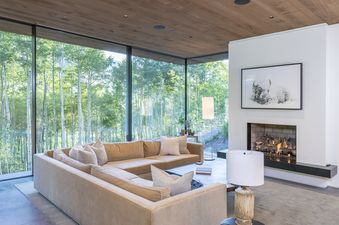 A living room with a large sectional sofa facing a window with a view of a forest. A fireplace is located to the right, with a black framed artwork depicting a bison above it.