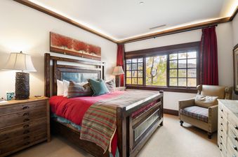 A rustic yet elegant master bedroom in Arrowleaf Lodge 211 features a large wooden bed with red bedding, a bedside lamp, an armchair by a window with mountain views, and a tree-themed artwork above the bed.