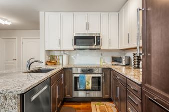 A modern kitchen with white upper cabinets, dark wood lower cabinets, and granite countertops features stainless steel appliances including a microwave, oven, and dishwasher, along with a sink.