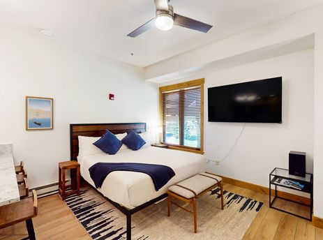 A cozy king-size bed with blue pillows and a navy throw blanket in EagleRidge Lodge 313's bedroom, featuring a flat-screen TV, a ceiling fan, and wooden blinds on the window.