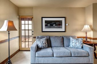 The living area of Copperbottom 205, featuring a gray sofa with patterned pillows, framed artwork, and lamps, with a door leading to a balcony.
