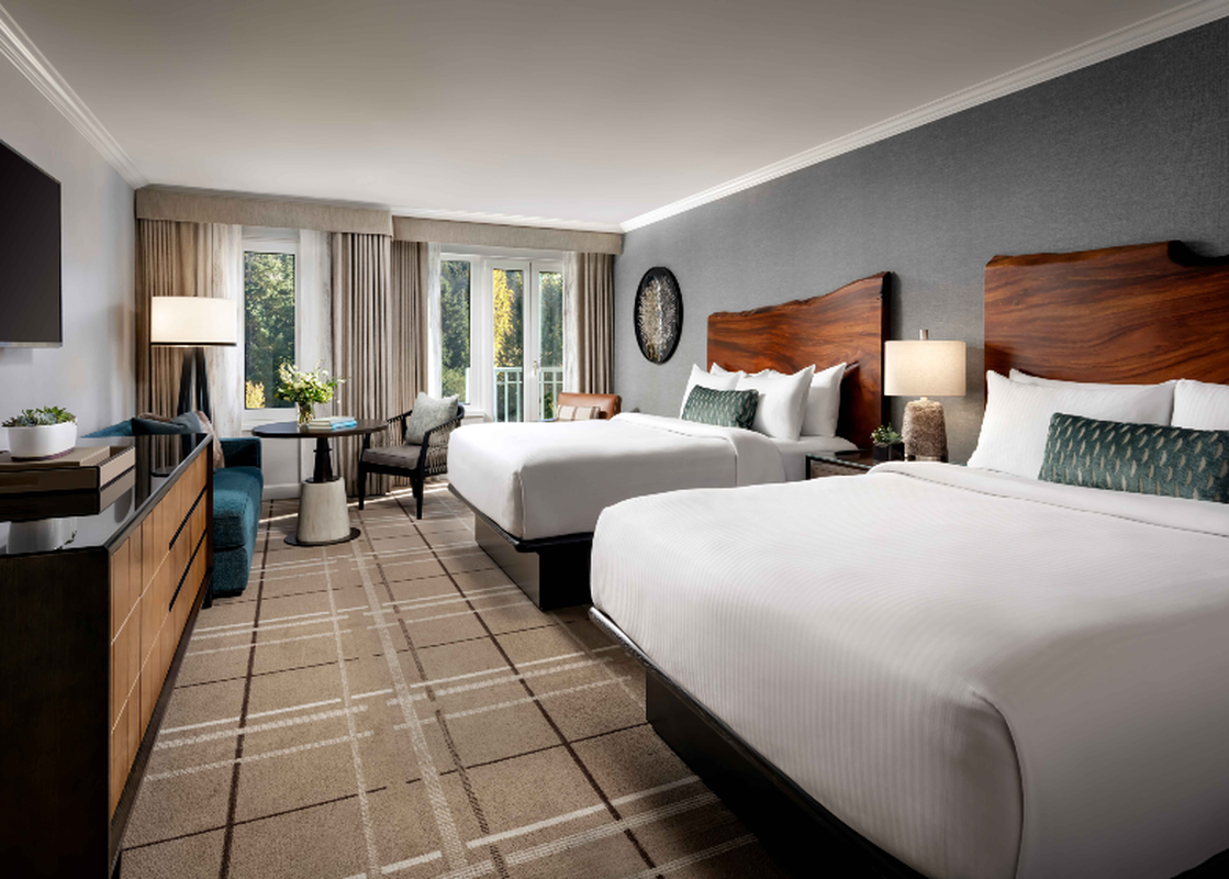 A spacious hotel room, the Fairmont Chateau Queen and Queen Room, features two queen-sized beds, a seating area with a small table, a dresser with a TV, and large windows looking out onto a lush green landscape.