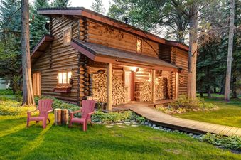 A rustic log cabin with a wooden porch and a walkway leading to the front door. Two red chairs are placed on the green lawn in front of the cabin.