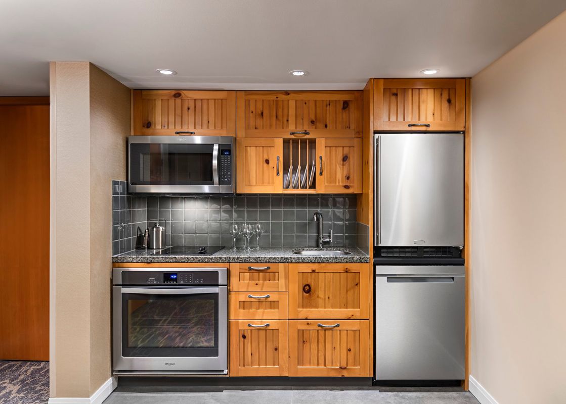 A fully equipped compact kitchen featuring wooden cabinetry, stainless steel appliances, and a grey tiled backsplash.