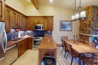 A kitchen with stainless steel appliances, wooden cabinets, and a granite countertop, open to a dining room with a wooden table and chairs, with a fireplace in the background.