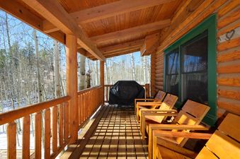 A covered wooden porch extends from a log cabin, featuring several wooden chairs and a black covered BBQ grill, all overlooking a snowy forest of tall, slender trees.