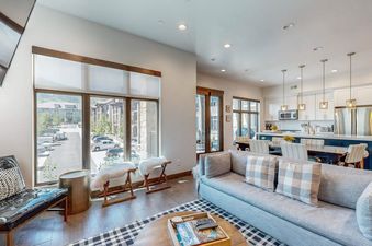 A modern open-concept living room with a gray sofa, a checkered rug, and large windows offering views of the outdoors, seamlessly connecting to a contemporary kitchen and dining area.