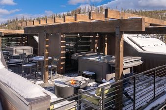 A snowy rooftop deck at the Starview Chalet featuring a hot tub, a lit fire pit, and patio furniture under a wooden pergola, with a mountain backdrop.