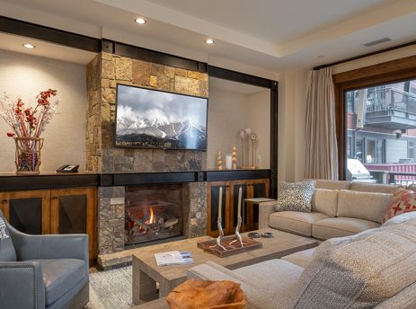 A cozy living room in Buck Mountain 2 Bedroom One Steamboat Place 307 features a stone fireplace, a television, comfortable seating, and a view of the snowy outdoors.