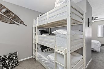 A bunk bed with white linens and a white wooden frame is shown in a bedroom. The bed is next to a wall with a gray paint finish. There is another white bed next to the bunk bed.