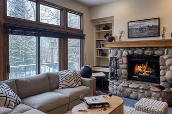 A cozy living room at Chalet Fortessa with a fireplace, comfortable sectional sofa, and large windows offering a snowy outdoor view.