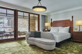 A cozy bedroom with a large wooden bed, a curved chaise lounge with pillows, and a large glass door leading to a snowy patio.