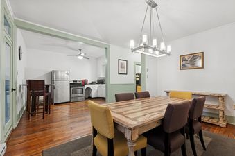 A wide shot of The Loft's open-concept dining room and kitchen, featuring a wooden dining table with mustard yellow and brown chairs, a contemporary chandelier, and a stainless steel kitchen setup.