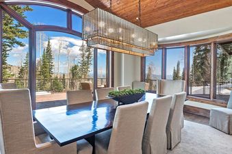 A luxurious dining room at Alpenglow at the Colony features a long, dark wood table with beige upholstered chairs, set against a backdrop of expansive windows showcasing stunning mountain and forest views.