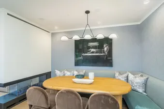 A modern dining area with a wooden oval table, velvet chairs, a built-in bench, and a modern chandelier, featuring a fireplace and large artwork on the wall.