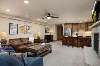 An inviting open-concept living area in Montaneros 220 features a leather sofa, blue armchairs, a wooden coffee table, a wet bar with stools, and a stone fireplace with a mounted television.