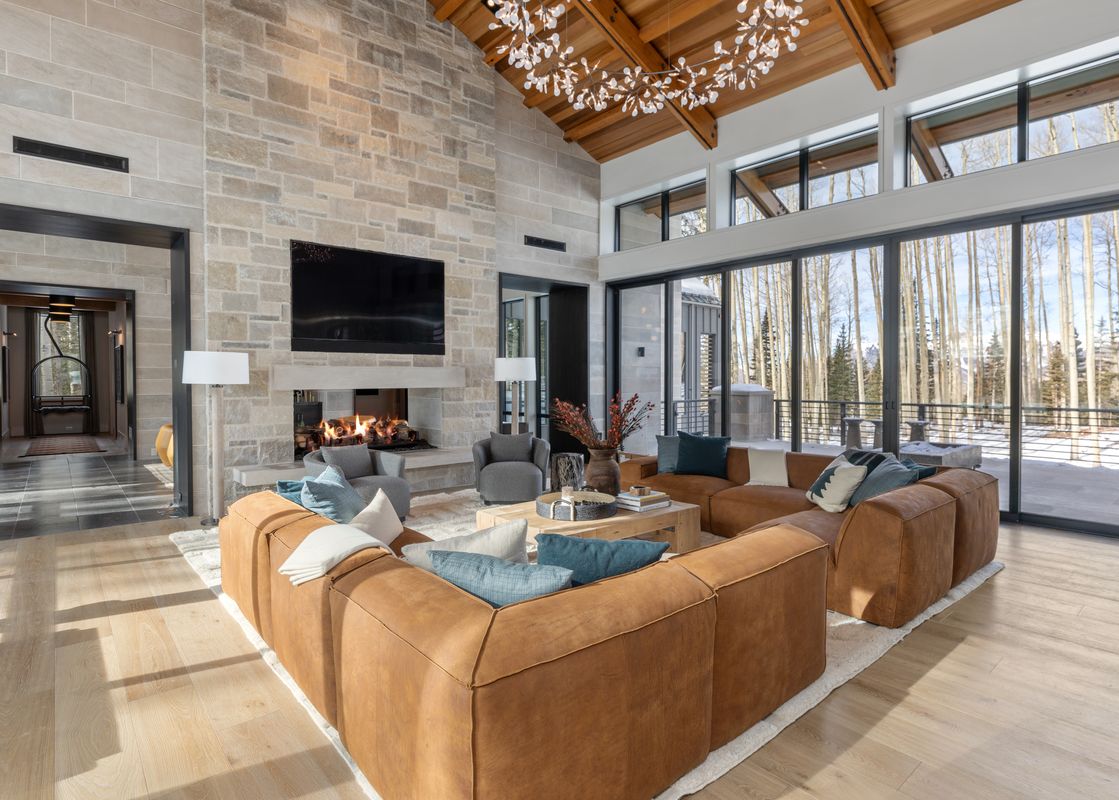 A spacious living room at Lone Star Lodge Mountain Village features a large brown sectional sofa, a stone fireplace, a flat-screen TV, and expansive windows offering a winter mountain view.