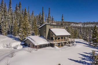 A modern mountain home, Lone Mountain Lookout, is nestled among snow-covered pine trees under a bright blue sky.