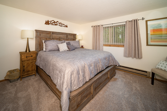 The primary bedroom features a large bed with a rustic wooden frame, light gray bedding, and various throw pillows, alongside a wooden nightstand with a lamp.