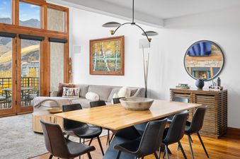 A modern dining area and living room with a large wooden table, black chairs, and a comfortable sectional sofa, featuring a balcony with mountain views and a decorative mirror reflecting a fireplace.