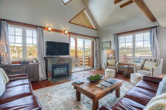 A cozy living room at Saddle Ridge Townhome Unit B1 with leather sofas, a fireplace, and large windows offering a panoramic view of snow-capped mountains.