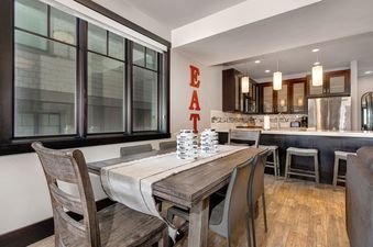 A dining room with a large wooden table and six chairs, a kitchen with a stainless steel refrigerator and a white countertop, and a window with a view of a building outside.