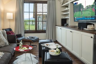 A comfortable lounge area with a large TV displaying golf, a sofa, a leather lounge chair, a red coffee table, and a large window overlooking a green landscape.
