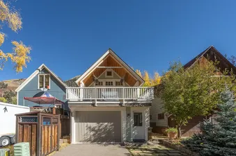 The exterior of PACIFIC HOUSE showcases a light green facade with wooden gables, a balcony, and a garage, surrounded by autumn trees under a clear blue sky.