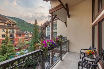 The Montaneros 402 balcony is a private outdoor space with two chairs, a table, and flower boxes, overlooking green mountains and nearby buildings.