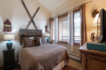 The master bedroom features a queen-sized bed, two nightstands with lamps, a dresser with a television, and windows with blinds and curtains.