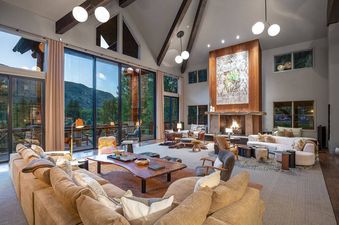A wide shot of a spacious and luxurious living room at Riverbend House, featuring large windows with mountain views, a grand fireplace, and multiple comfortable seating areas.