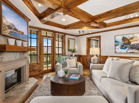 A cozy and elegant living room at Chateau Beaver Creek Residence 309, featuring a stone fireplace, wood-beamed ceiling, comfortable seating, and large windows with an outdoor view.