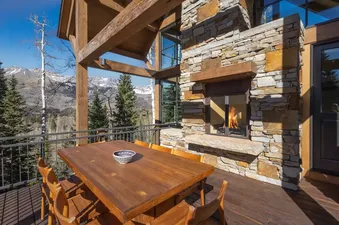 An outdoor wooden dining deck at Galloping Goose Chalet featuring a large stone fireplace with a burning fire and scenic mountain views.