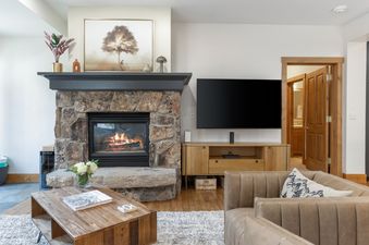 A cozy living room at 118 Villa Montane features a stone fireplace with a warm fire, a television mounted above a wooden media console, and comfortable seating.