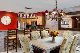 A dining table with six paisley upholstered chairs and a floral centerpiece, connected to a kitchen with wooden cabinets and stainless steel appliances.