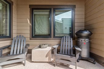 A private balcony at Central Loft 303 features two Adirondack chairs and a modern concrete side table, with a black Fuego grill positioned to the right against the building's light brown siding.
