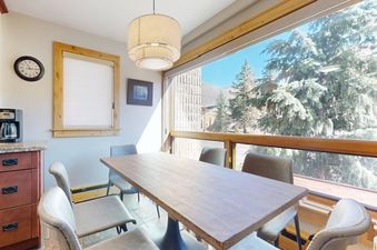 The dining area at Vail 21 303 showcases a wooden table, upholstered chairs, a coffee maker, and large windows revealing a scenic outdoor view with pine trees and mountains.